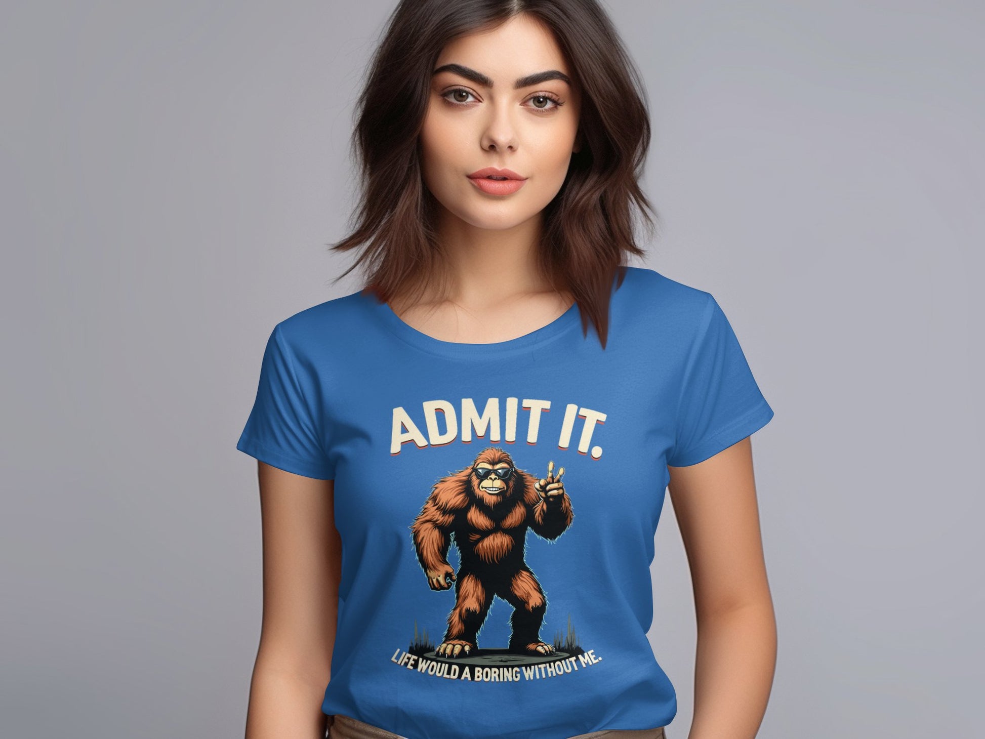 A brown-haired woman wears a Garment Graphics blue T-shirt featuring the playful Admit It Bigfoot design, with a Bigfoot illustration and the text, ADMIT IT. Life would be boring without me, all set against a plain gray backdrop.