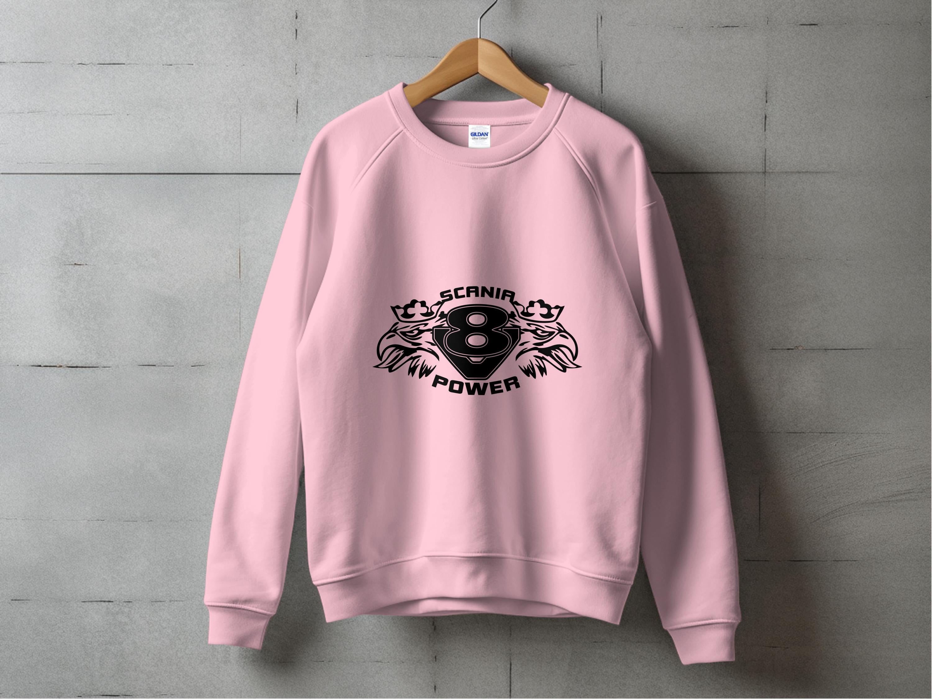 A pink sweatshirt from Garment Graphics hangs on a wooden hanger against a gray wall, featuring the Scania Power 8 logo with two lions flanking the number eight and SCANIA POWER boldly printed in black.