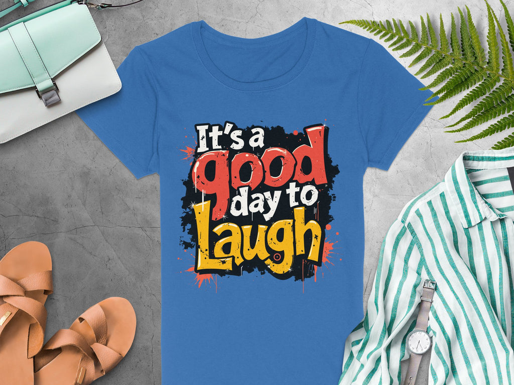 A Garment Graphics semi-fitted blue t-shirt features a bold, colorful Its a good day to laugh graphic. Displayed on gray fabric, its surrounded by a green striped shirt, watch, tan sandals, handbag, and fern leaf, all enhancing the shirts cheerful vibe.