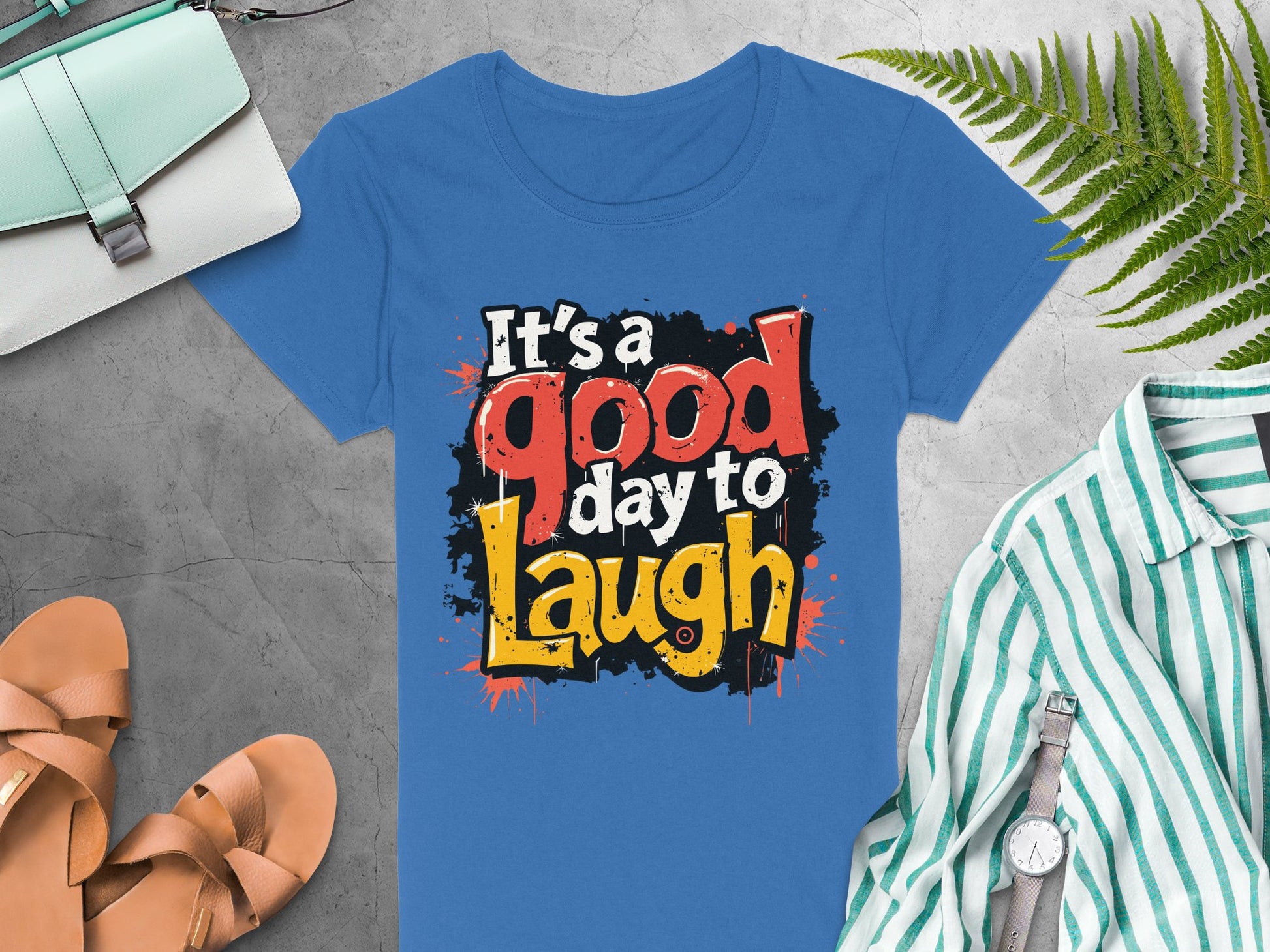A Garment Graphics semi-fitted blue t-shirt features a bold, colorful Its a good day to laugh graphic. Displayed on gray fabric, its surrounded by a green striped shirt, watch, tan sandals, handbag, and fern leaf, all enhancing the shirts cheerful vibe.