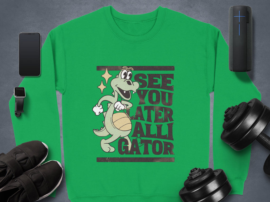 A Garment Graphics green sweatshirt showcases a fun alligator graphic and the playful text See You Later Alligator, surrounded by a smartphone, smartwatch, earphones, dumbbells, speakers, and black shoes.