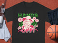 A Garment Graphics black classic fit T-shirt rests on a basketball court, featuring a cowboy hat design with flowers and a lollipop. It boldly states HANDS OFF! adorned with small hearts. Nearby essentials include a basketball, shorts, towel, and water bottle.