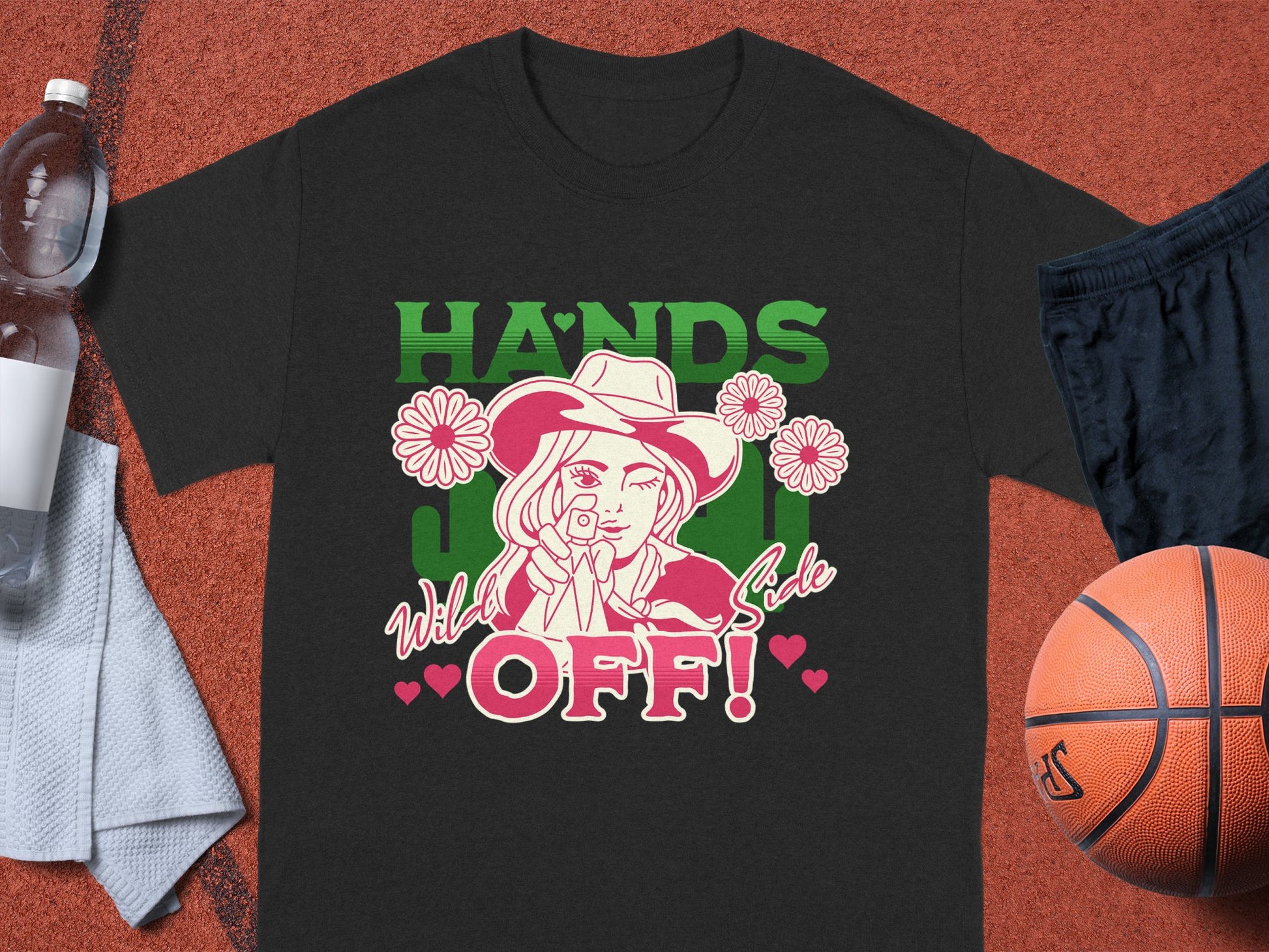 A Garment Graphics black classic fit T-shirt rests on a basketball court, featuring a cowboy hat design with flowers and a lollipop. It boldly states HANDS OFF! adorned with small hearts. Nearby essentials include a basketball, shorts, towel, and water bottle.