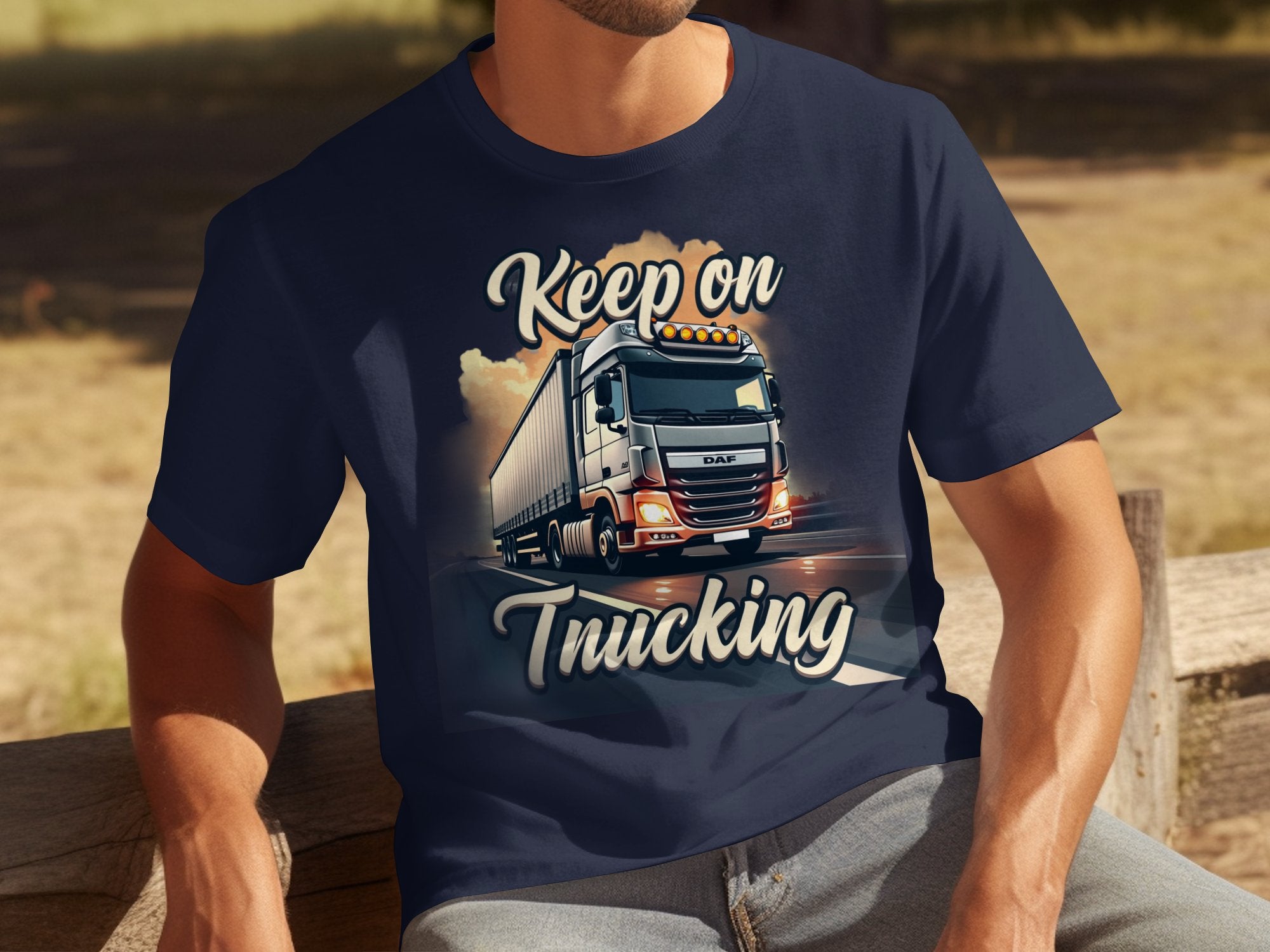 In a sunny outdoor area with lush trees and green grass, someone wears a navy blue Garment Graphics t-shirt featuring a semi-truck graphic and the words Keep on Trucking while relaxing on a wooden bench. Ideal for truck enthusiasts.