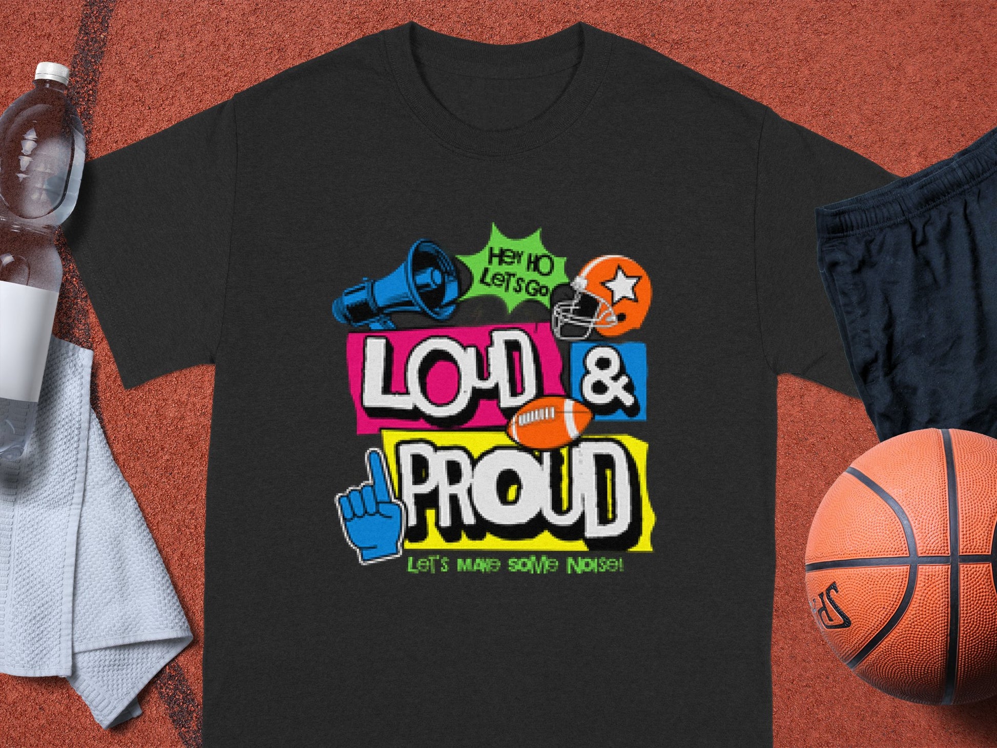 Garment Graphics presents a black T-shirt in family sizes featuring vibrant LOUD & PROUD text with sports graphics including a helmet, football, megaphone, and foam finger. Nearby are a basketball, shorts, towel, and water bottle on the red athletic court.