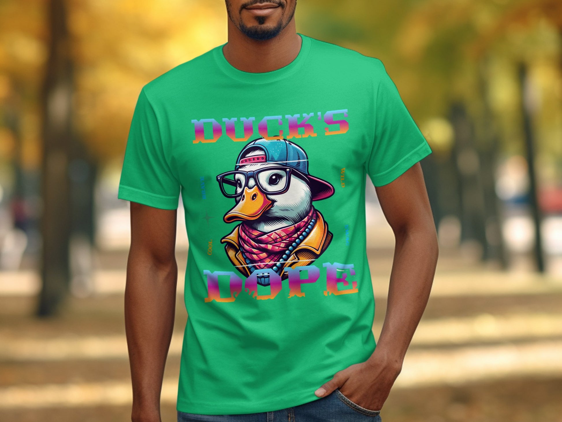 A man wears a bright green T-shirt from Garment Graphics featuring a cartoon duck with a cap, glasses, and scarf. The words Ducks Dope are printed above and below the design, making it pop against the backdrop of trees with vivid yellow and orange leaves.