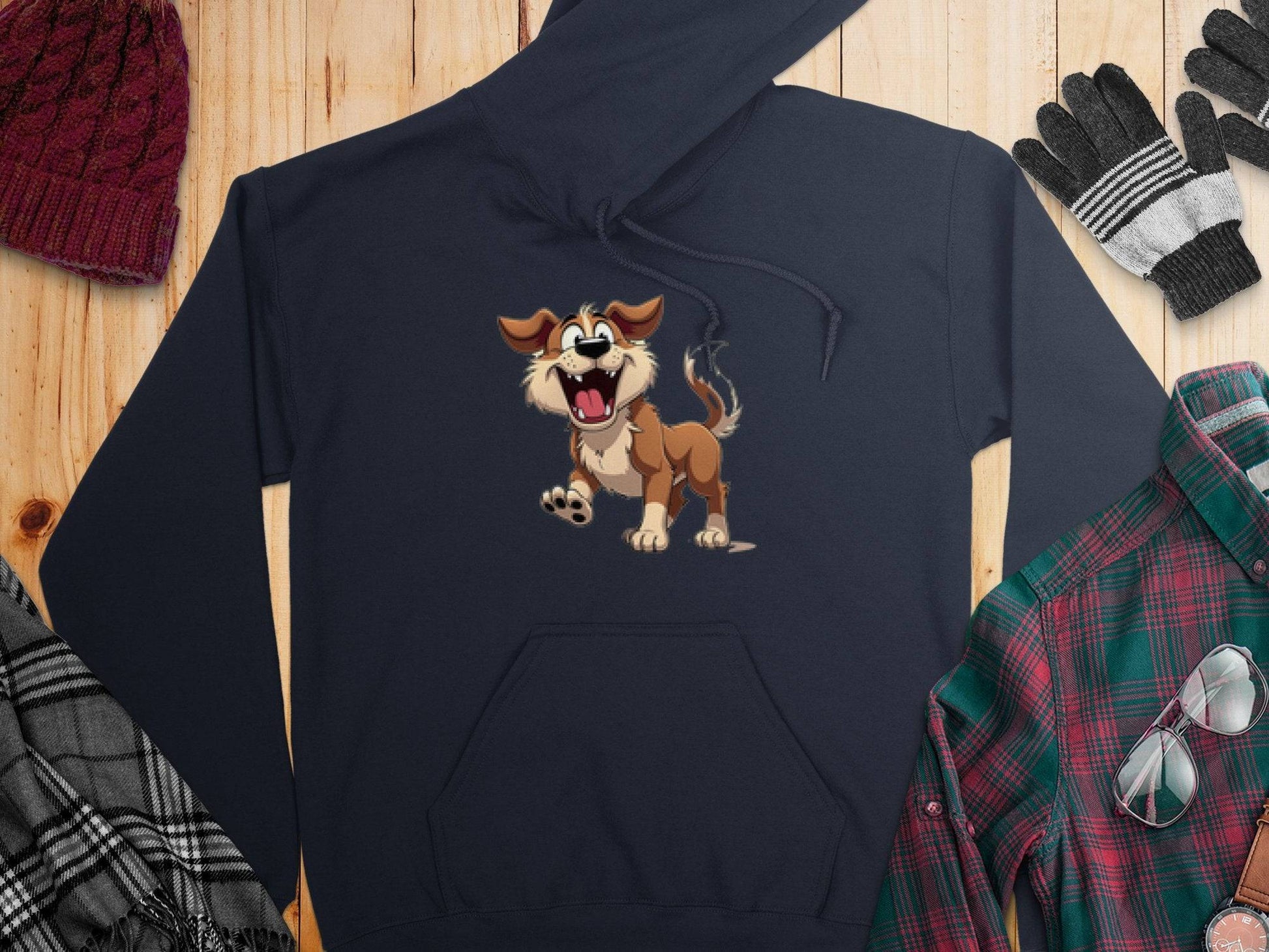 A navy cartoon puppy hoodie by Garment Graphics, perfect for dog lovers, rests on a wooden surface. Its surrounded by a red knit hat, striped gloves, a gray checkered scarf, and a green plaid shirt topped with sunglasses.