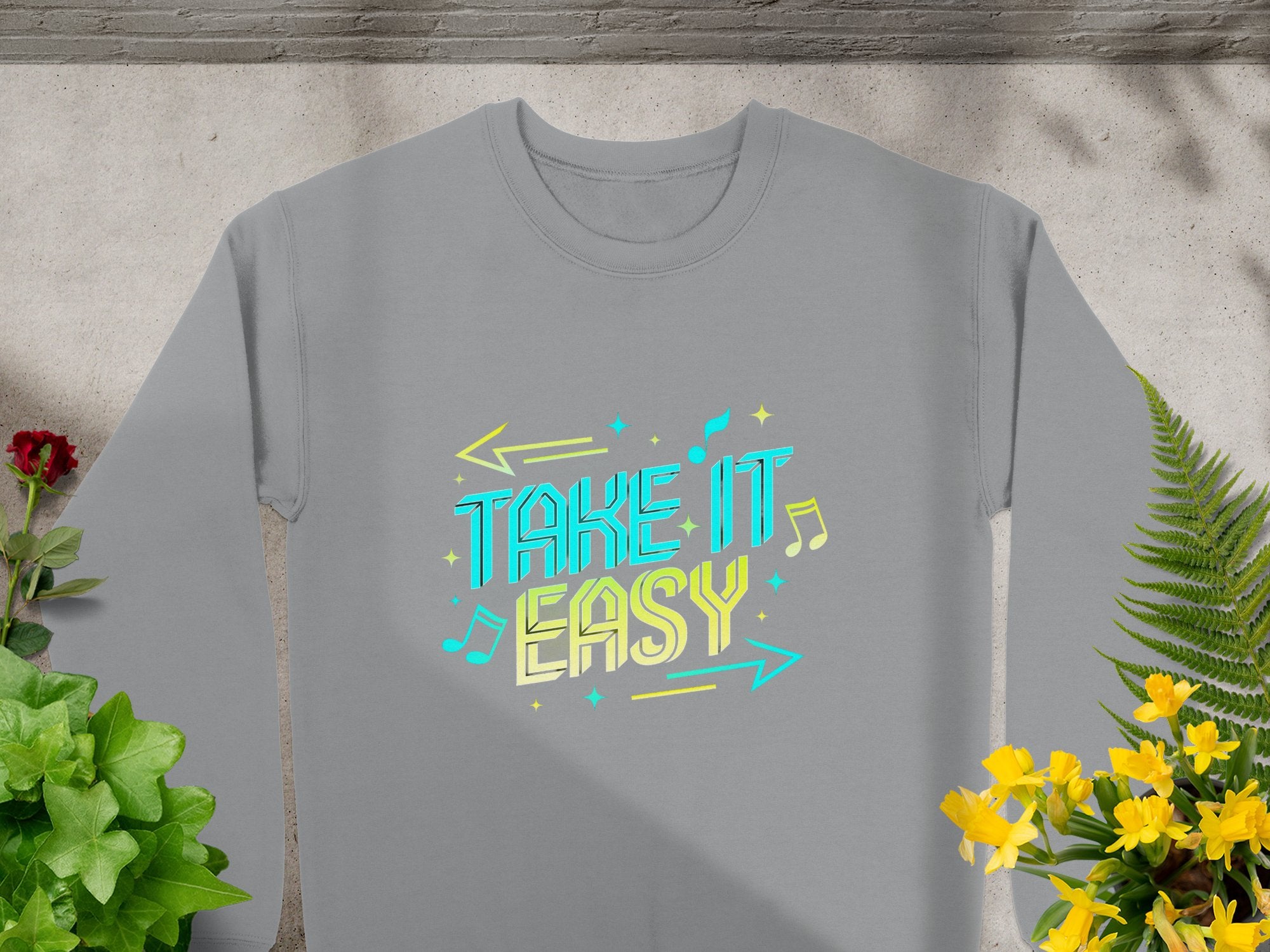 Garment Graphics presents a unisex sweatshirt, showcasing the Take It Easy design in blue and yellow with arrow and music note accents. Set among greenery on concrete, its ideal for casual wear.