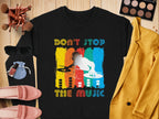 The Garment Graphics black DJ design T-shirt with a silhouette and colorful stripes embodies music lovers spirit. The Dont Stop the Music text pops, complemented by a yellow blazer, pink shoes, sunglasses, makeup brushes, and cosmetics on a wooden surface.