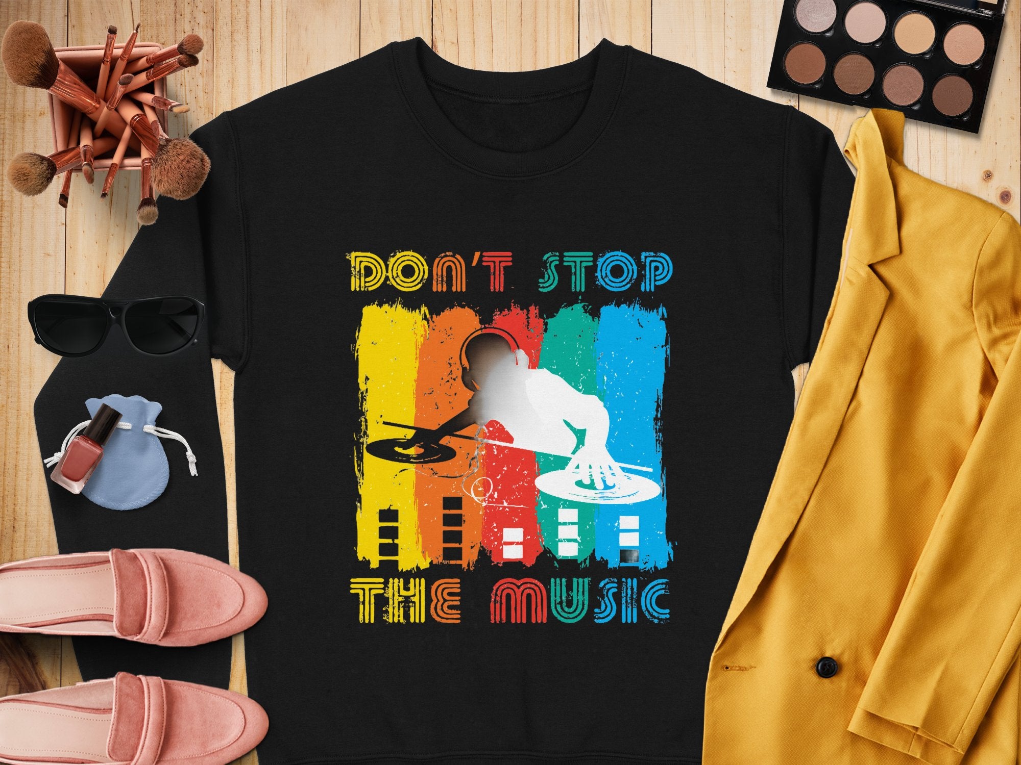 The Garment Graphics black DJ design T-shirt with a silhouette and colorful stripes embodies music lovers spirit. The Dont Stop the Music text pops, complemented by a yellow blazer, pink shoes, sunglasses, makeup brushes, and cosmetics on a wooden surface.