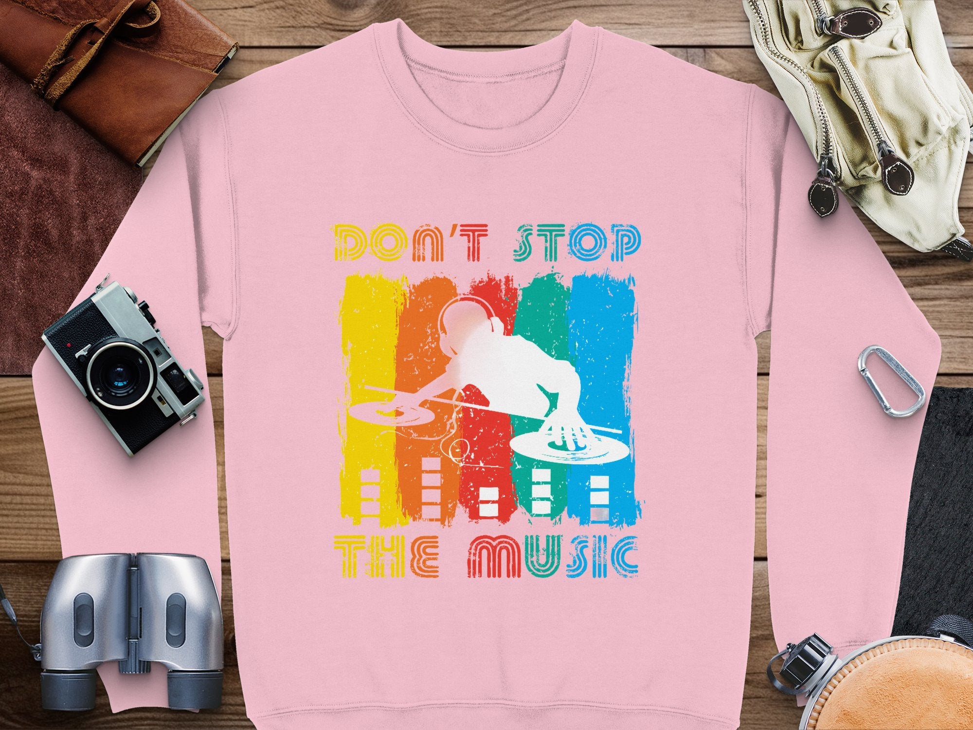 A pink Garment Graphics sweatshirt with a vibrant DJ design and Dont Stop the Music slogan lies on a wooden surface, ideal for music lovers. Nearby items include a camera, binoculars, leather bag, notebook, and carabiner clip.