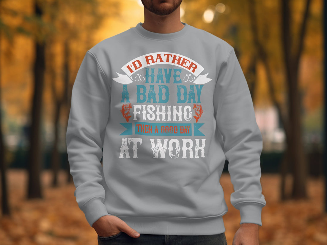 A white Garment Graphics t-shirt with Id rather have a bad day fishing than a good day at work sits on a plaid shirt. Nearby, headphones and a camera lie on the wooden surface—ideal for fishing fans who prefer unisex sizing in their casual wear.