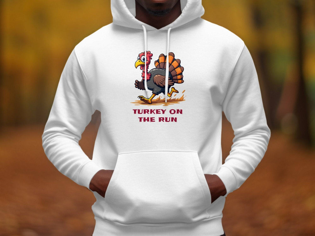 Garment Graphics black hoodie features a cartoon turkey in running shoes with humorous Turkey On The Run text. Pine cones, ornaments, and a cozy coffee cup complement the festive wooden background, making it a quirky hoodie to cherish.