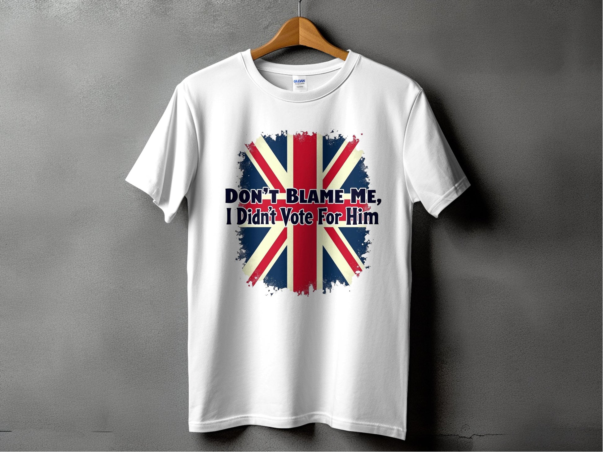 Garment Graphics offers a white classic fit T-shirt featuring a distressed Union Jack design. The bold statement DONT BLAME ME, I Didnt Vote For Him is displayed in prominent text. The shirt is shown on a wooden hanger against a gray backdrop.