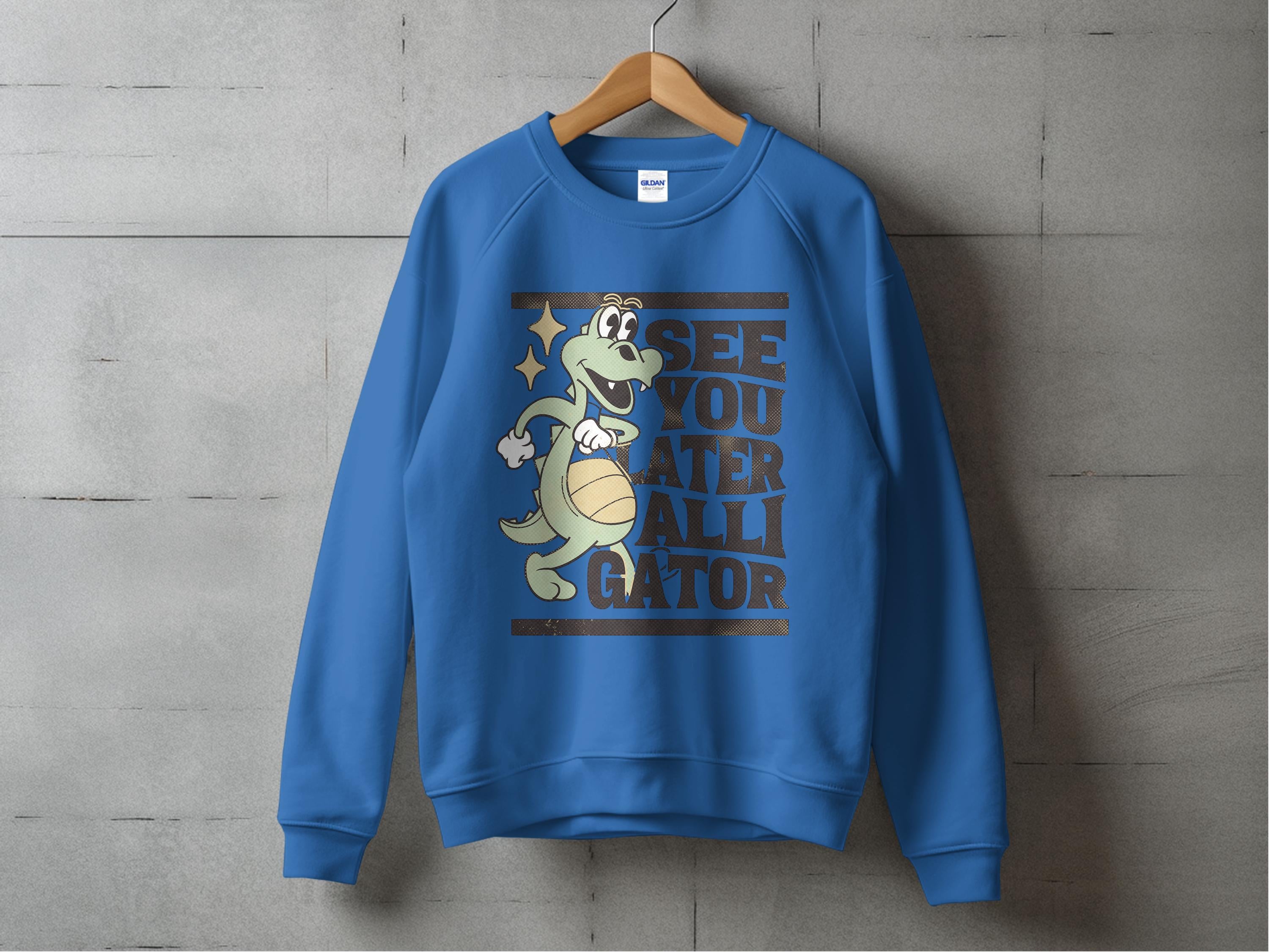 A Garment Graphics sweatshirt with a cartoon alligator holding a basketball hangs on a concrete wall, featuring a speech bubble that says, See you later alligator.