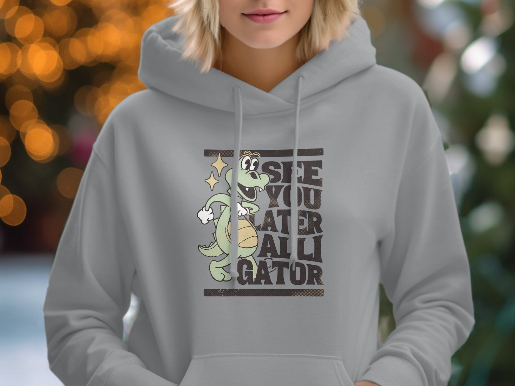 A person is wearing a classic fit hoodie by Garment Graphics featuring a cartoon alligator holding a basketball. The background is softly blurred with twinkling lights.