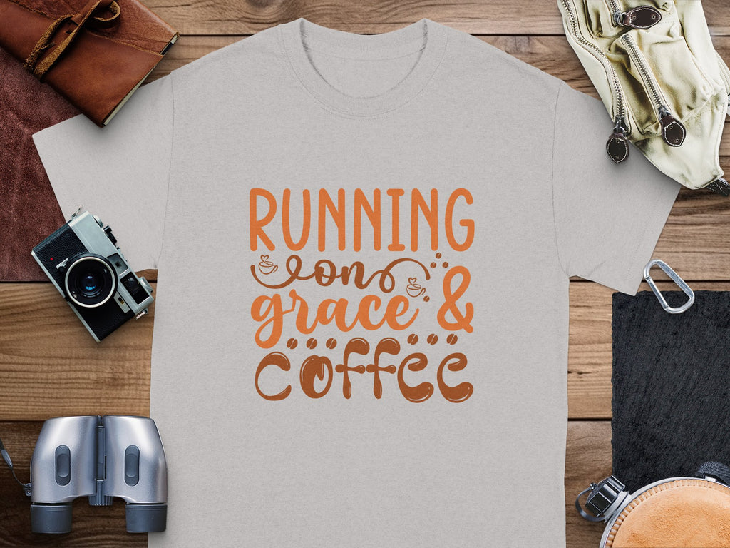 A light gray Garment Graphics t-shirt with Running on Grace & Coffee in orange and brown script lies on a wooden surface, surrounded by essentials like a wallet, camera, binoculars, notebook, and glasses.