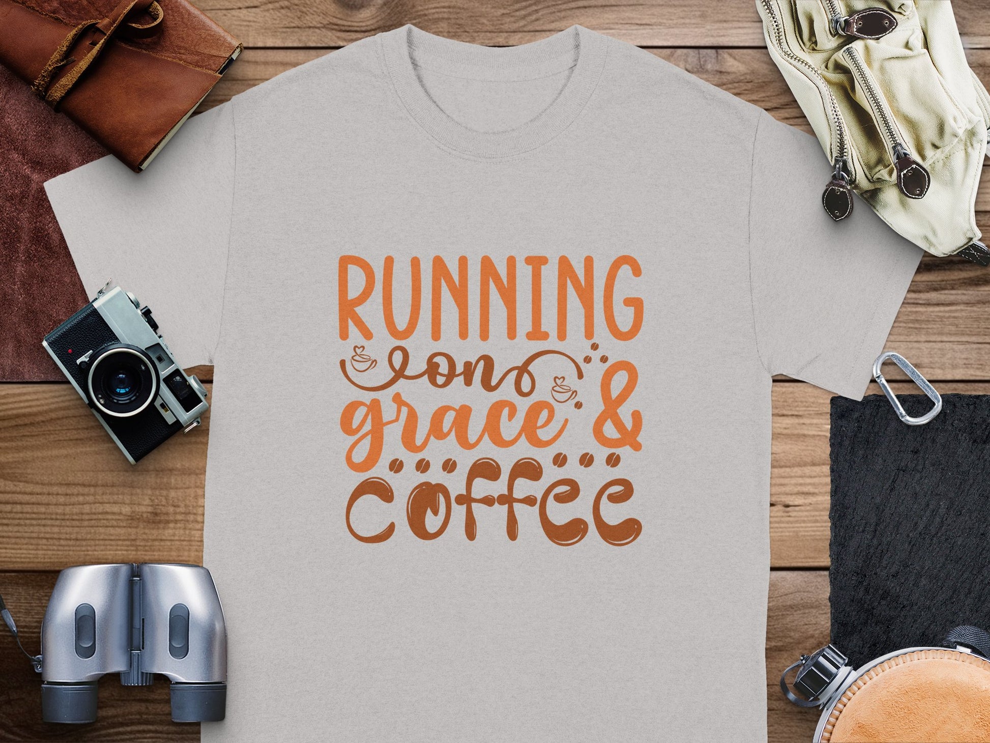 A light gray Garment Graphics t-shirt with Running on Grace & Coffee in orange and brown script lies on a wooden surface, surrounded by essentials like a wallet, camera, binoculars, notebook, and glasses.