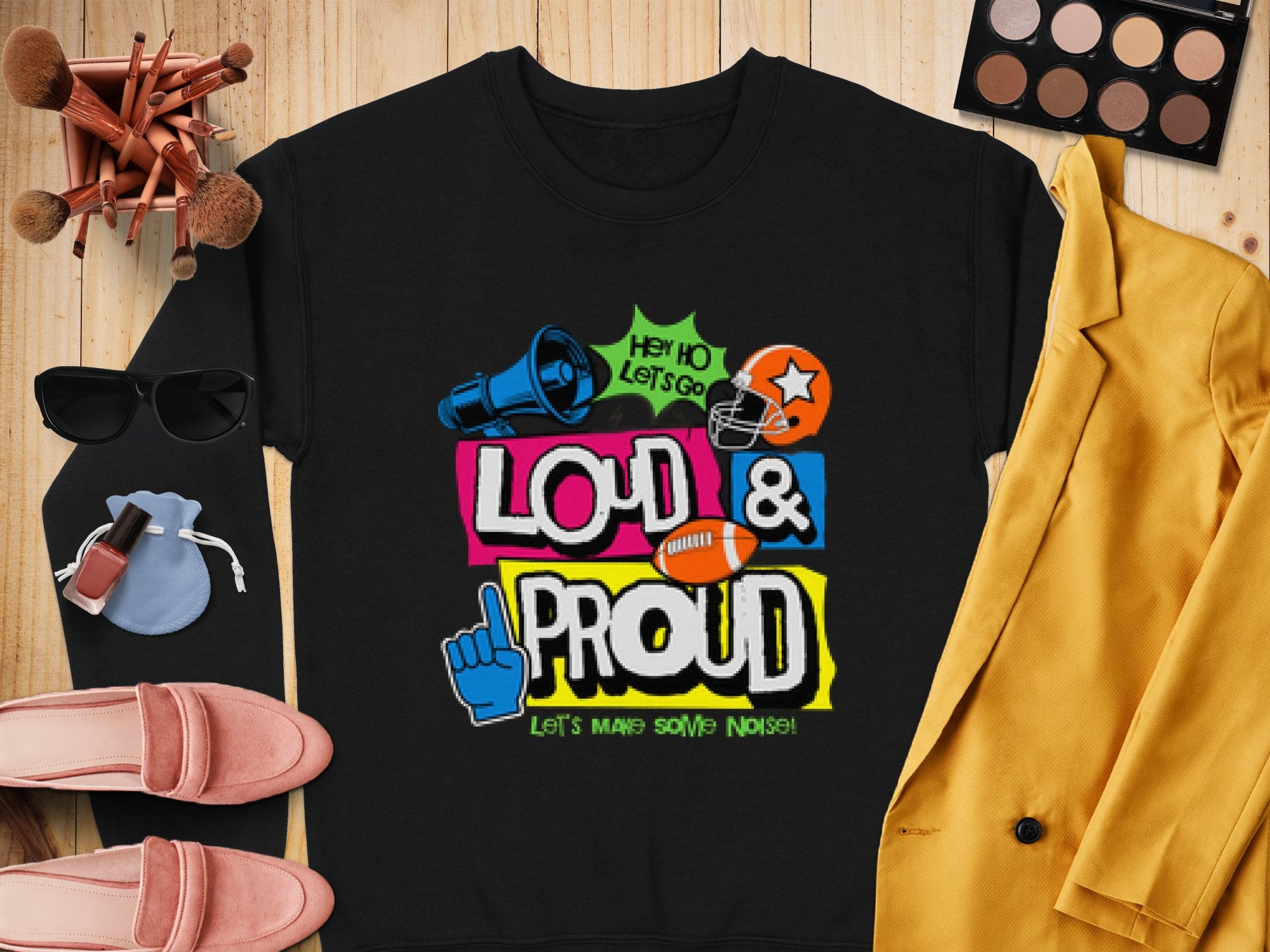 Garment Graphics presents a unique black unisex shirt with vibrant text, Hey Ho Lets Go Loud & Proud Lets Make Some Noise! accompanied by symbols like a megaphone, football helmet, and foam finger. Styled on wood with cosmetics, a yellow blazer, and pink shoes for added flair.