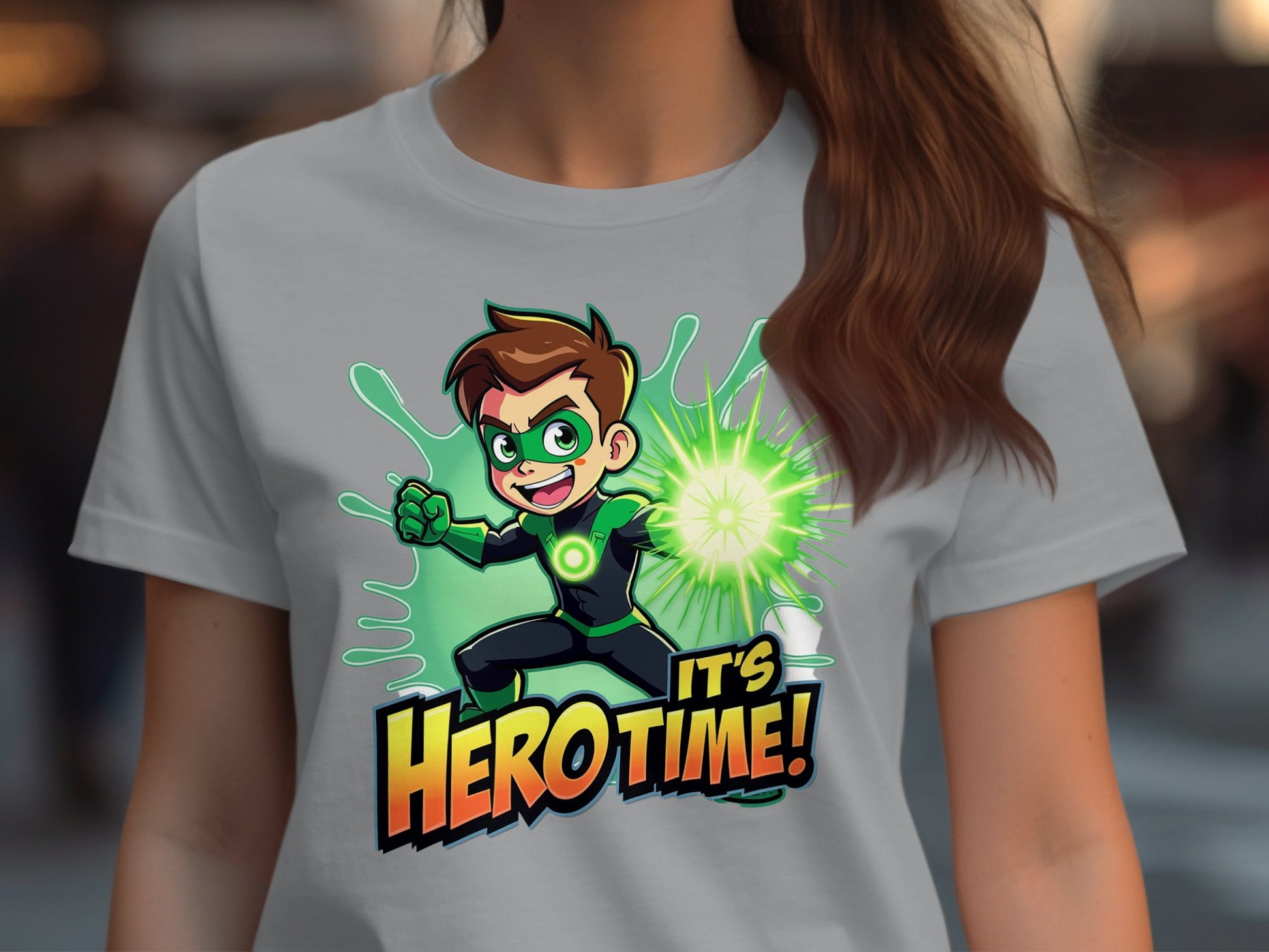 A person wears a light gray Garment Graphics t-shirt with a vibrant superhero design, featuring a cartoon hero with brown hair, green mask and suit, emitting a green energy blast. The words Its Hero Time! complete this perfect addition for young heroes at heart.