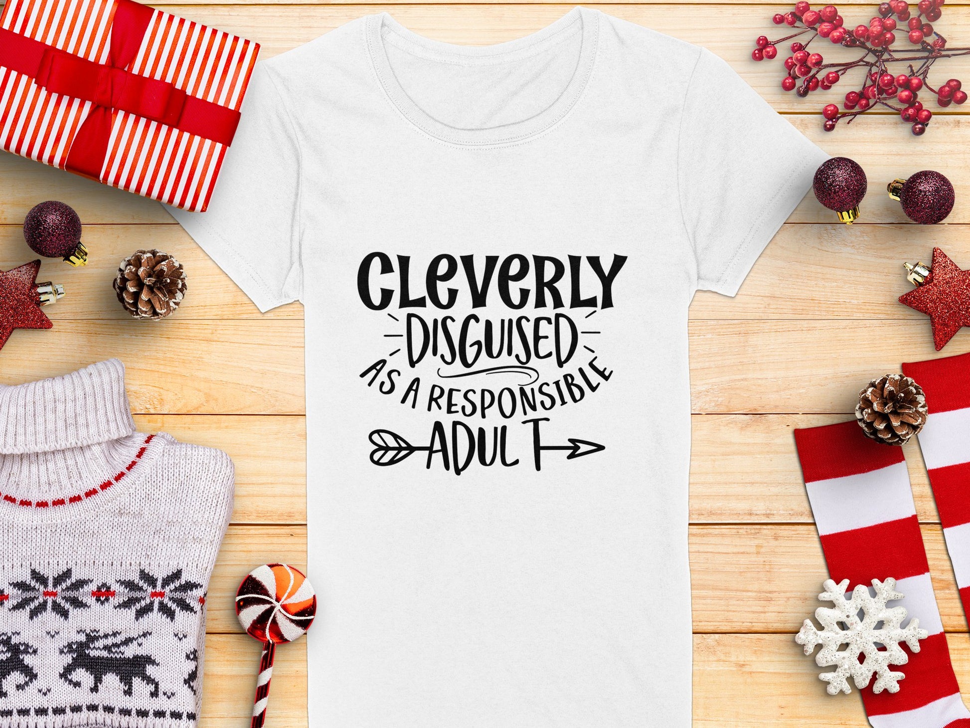 A semi-fitted white Garment Graphics T-shirt with Cleverly Disguised as a Responsible Adult is centered on a wooden surface, surrounded by holiday decor such as wrapped gifts, pine cones, a cozy sweater, candy cane, and festive decorations.