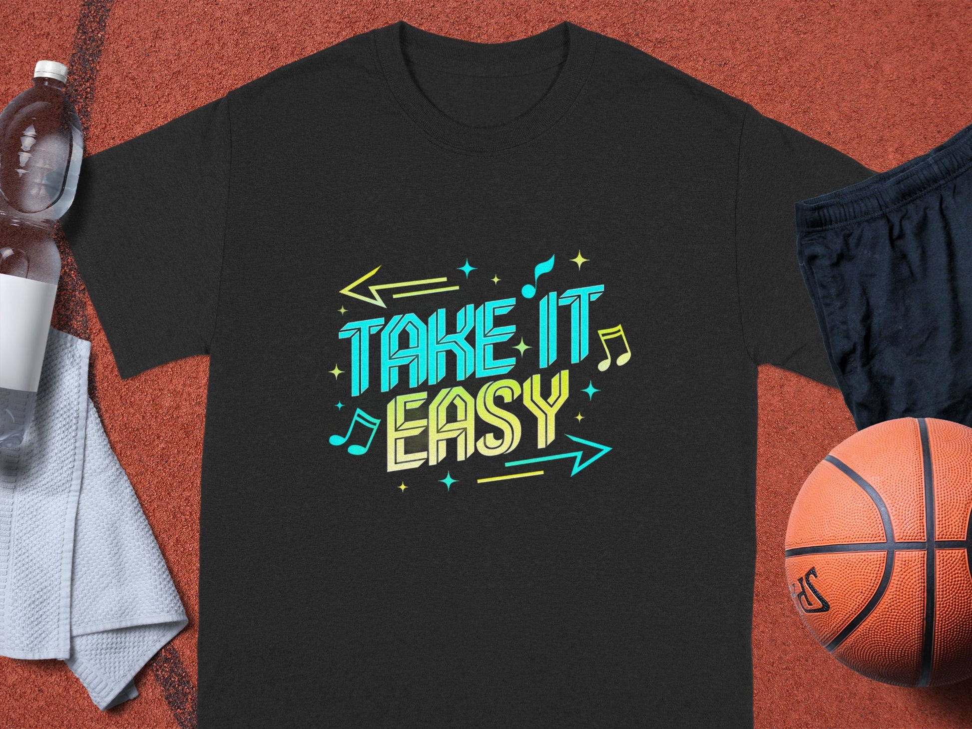 A Garment Graphics dark Take It Easy T-shirt showcases vibrant blue and yellow stylized text surrounded by a basketball, black shorts, a white towel, and a clear water bottle on a reddish-brown surface, capturing the rhythm of musical notes in motion.