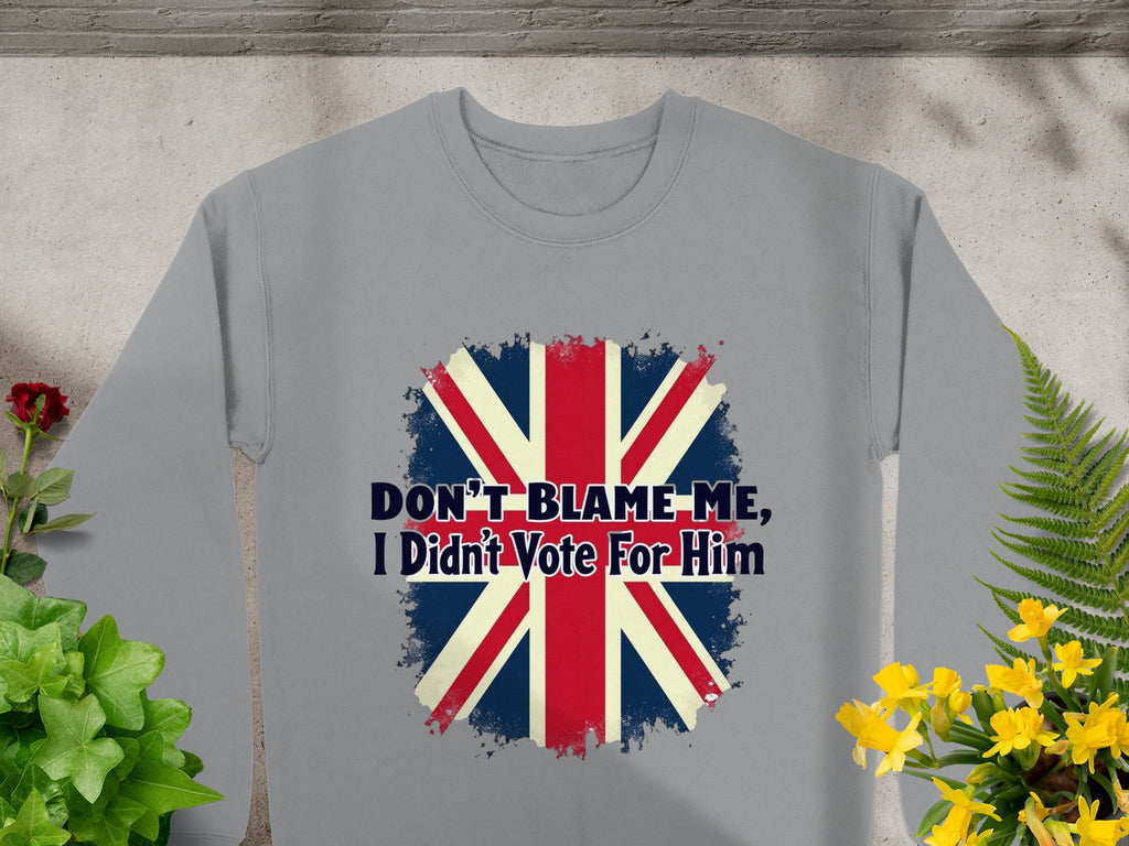 A gray Garment Graphics sweatshirt with a Union Jack and Dont Blame Me, I Didnt Vote For Him lies flat among green and yellow potted plants on a textured surface. This unisex piece adds a bold statement to any wardrobe.