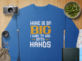 A Garment Graphics sweatshirt, featuring the bold text MINE IS SO BIG I HAVE TO USE BOTH HANDS, makes a statement as it rests on a wooden surface next to a camera, lens, kiwi slices in a bowl, a small map, book, and drawstring pouch.