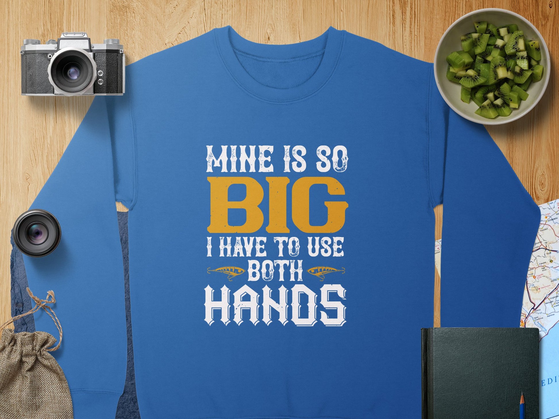 A Garment Graphics sweatshirt, featuring the bold text MINE IS SO BIG I HAVE TO USE BOTH HANDS, makes a statement as it rests on a wooden surface next to a camera, lens, kiwi slices in a bowl, a small map, book, and drawstring pouch.
