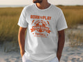 A person wears a Garment Graphics white T-shirt featuring bold orange text, Born to Play: One Goal One Dream, with Young Squad Football and a soccer ball graphic. It reads Kick. Score. Celebrate! over a vibrant grassy background.