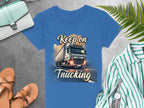A blue T-shirt from Garment Graphics features a truck design with Keep on Trucking. Its styled on a gray surface with a green-striped shirt, watch, brown sandals, mint purse, and green fern leaves.