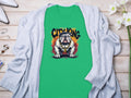 A green T-shirt by Garment Graphics features a bulldog with City King above, offering a unique style. It rests on wood, partially covered by a light gray cardigan, with white shoes, jewelry, and delicate white flowers nearby.