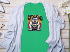 A green T-shirt by Garment Graphics features a bulldog with City King above, offering a unique style. It rests on wood, partially covered by a light gray cardigan, with white shoes, jewelry, and delicate white flowers nearby.