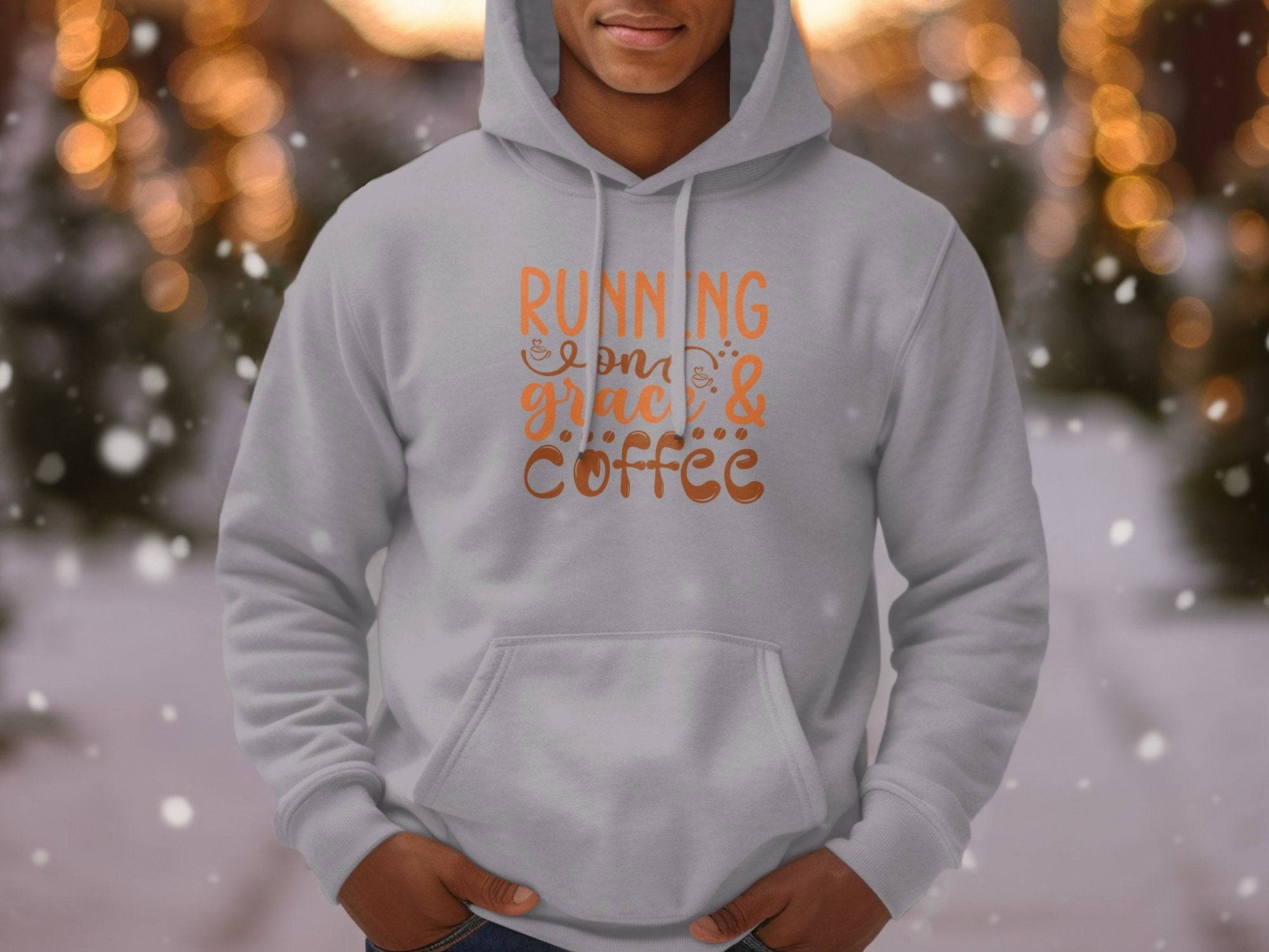 A person wears Garment Graphics classic fit Running on Grace and Coffee hoodie, showcasing orange and brown lettering on light gray medium-heavy fabric. The backdrop offers a blurred festive scene with twinkling lights and snowflakes.