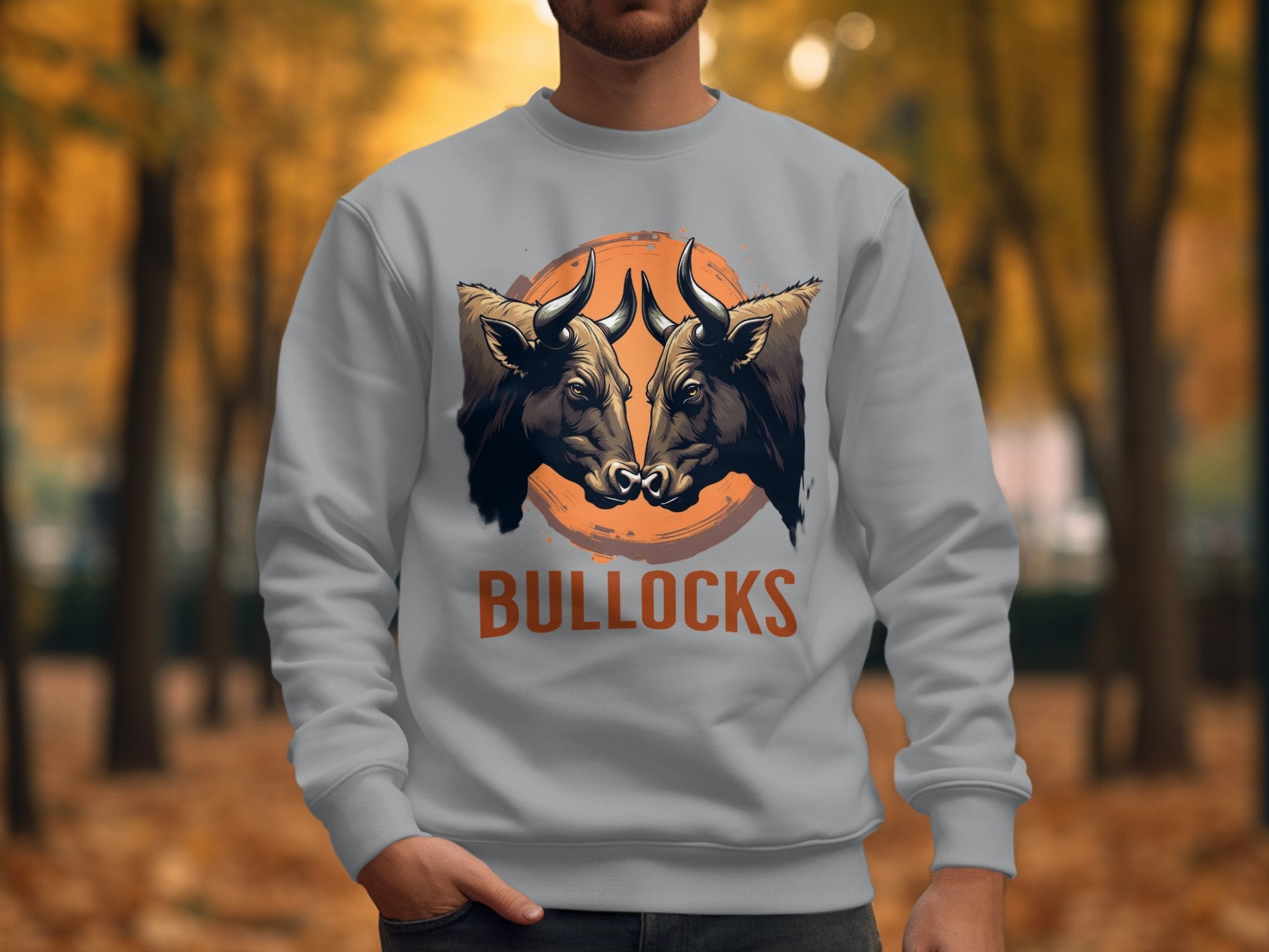 Someone wears a Garment Graphics bull graphic sweatshirt, showcasing two bull heads on an orange circle, with BULLOCKS boldly underneath. The backdrop is a forest with autumn colors, and the unisex sizing provides both versatility and style.