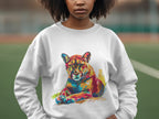 A woman poses outdoors in front of a blurred sports field, wearing a white Garment Graphics sweatshirt with a vibrant cougar design. With unisex sizing, this piece highlights her unique style.