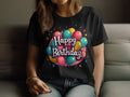 A person is seated on a couch wearing a vibrant Garment Graphics T-shirt made of 100% cotton, adorned with colorful balloons and Happy Birthday text. Only the torso and part of the face are visible, highlighting an indoor birthday celebration atmosphere.