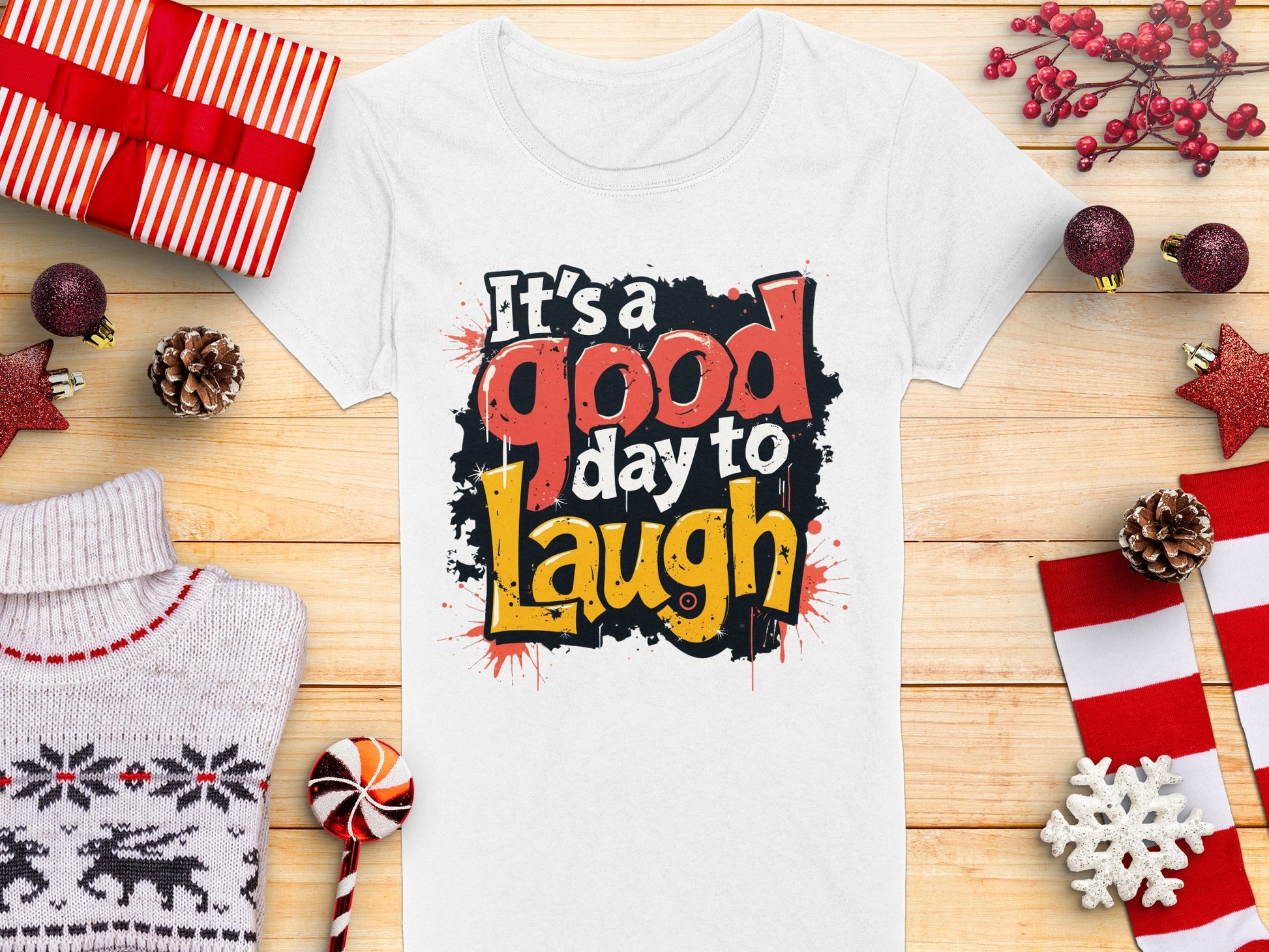 A semi-fitted white graphic print T-shirt from Garment Graphics, featuring Its a good day to Laugh in bold, colorful letters, is displayed on a wooden surface surrounded by wrapped gifts, pine cones, red ornaments, a cozy sweater, and a candy cane radiating positivity.
