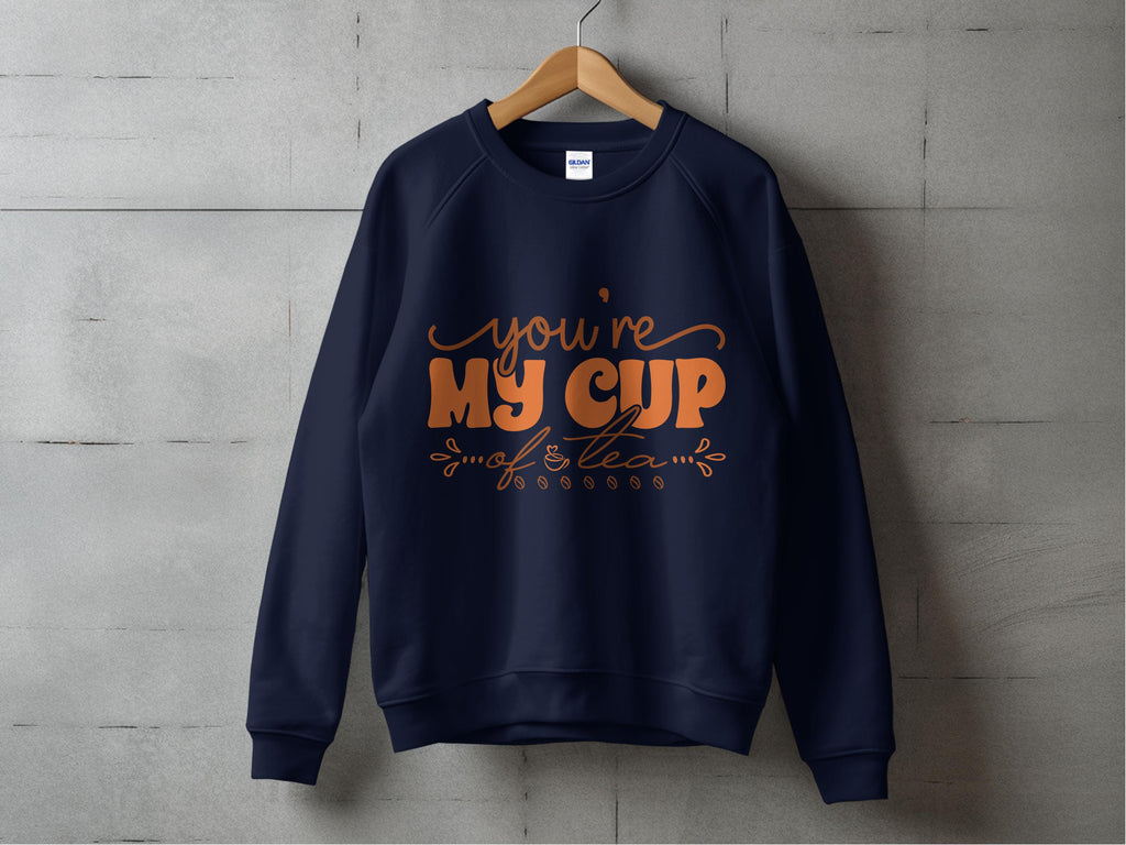 A Garment Graphics navy blue sweatshirt hangs on a wooden hanger against a concrete wall, radiating warmth for tea lovers. It features cozy orange text, Youre My Cup of Tea, complete with decorative flourishes.