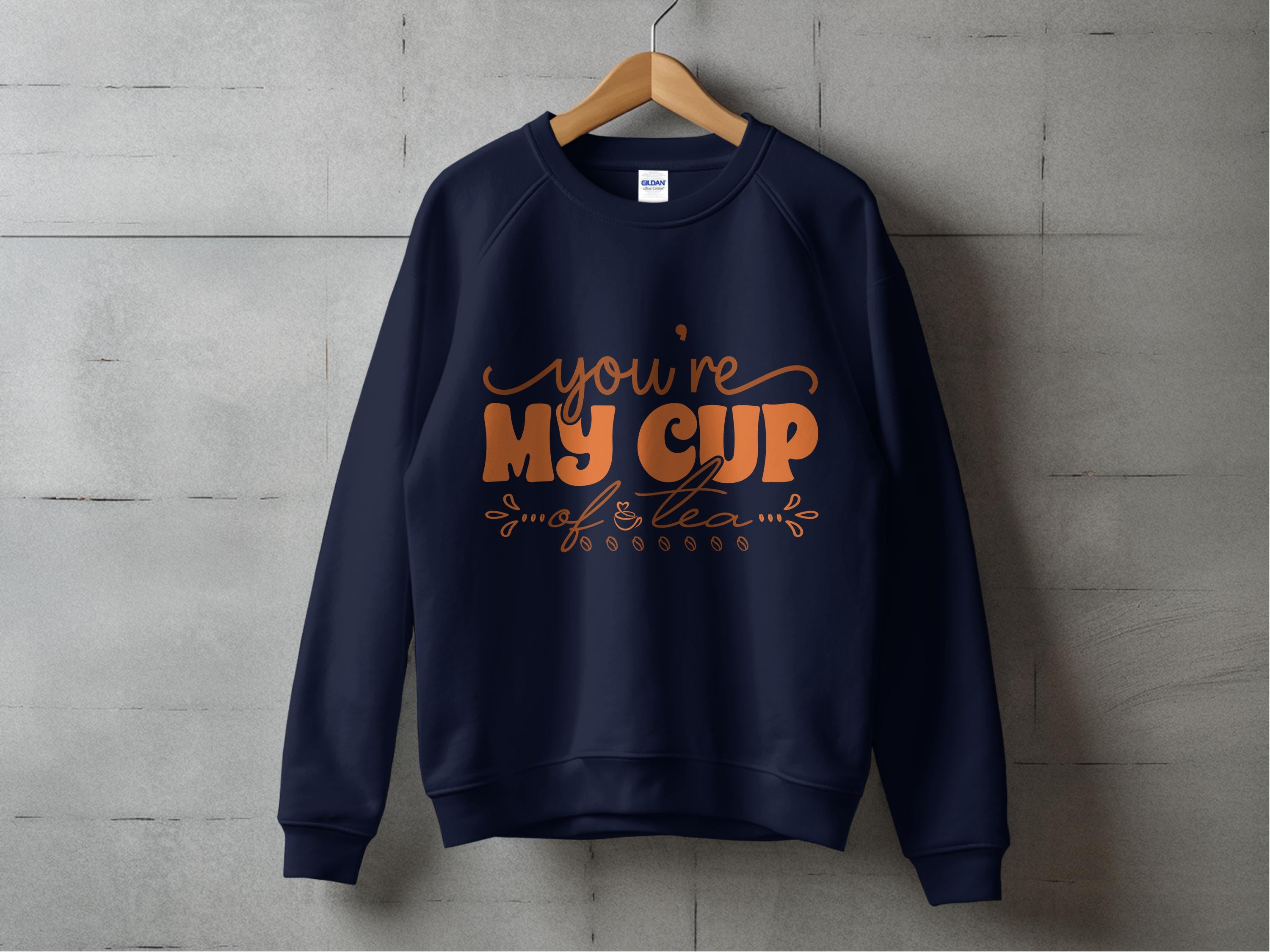 A Garment Graphics navy blue sweatshirt hangs on a wooden hanger against a concrete wall, radiating warmth for tea lovers. It features cozy orange text, Youre My Cup of Tea, complete with decorative flourishes.