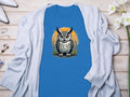 Garment Graphics blue T-shirt features a detailed owl graphic over an orange circle. Displayed on a wooden surface with a light gray cardigan, the shirt exudes artistic flair. Nearby, flowers and accessories complement the aesthetic.