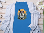 Garment Graphics blue T-shirt features a detailed owl graphic over an orange circle. Displayed on a wooden surface with a light gray cardigan, the shirt exudes artistic flair. Nearby, flowers and accessories complement the aesthetic.