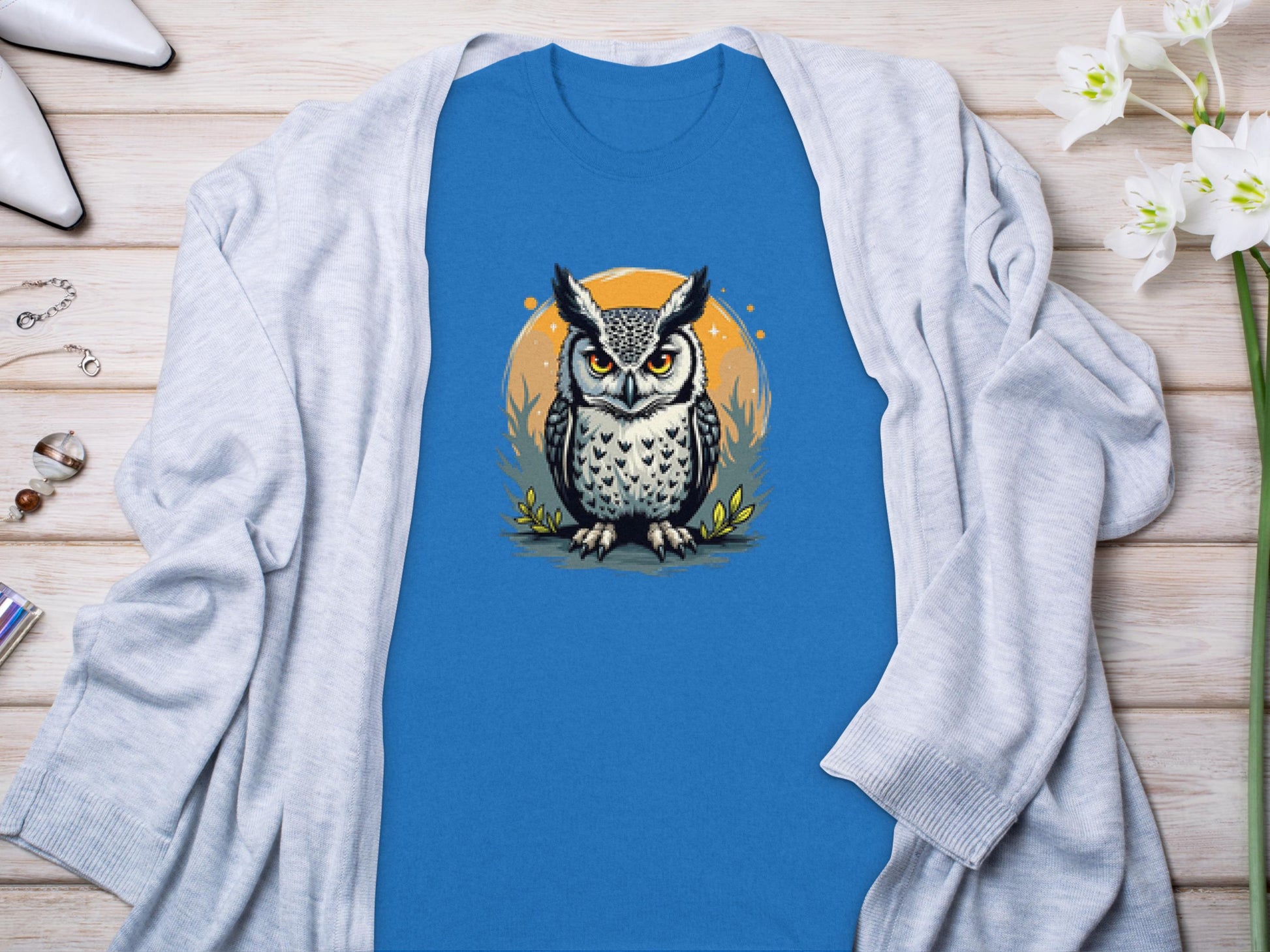 Garment Graphics blue T-shirt features a detailed owl graphic over an orange circle. Displayed on a wooden surface with a light gray cardigan, the shirt exudes artistic flair. Nearby, flowers and accessories complement the aesthetic.