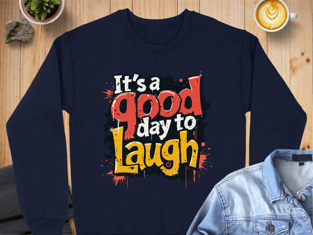 Garment Graphics offers a navy sweatshirt with Its a good day to Laugh in vibrant letters, featuring unisex sizing and medium-heavy fabric. Its styled with a denim jacket, a latte, and a small plant on wood.