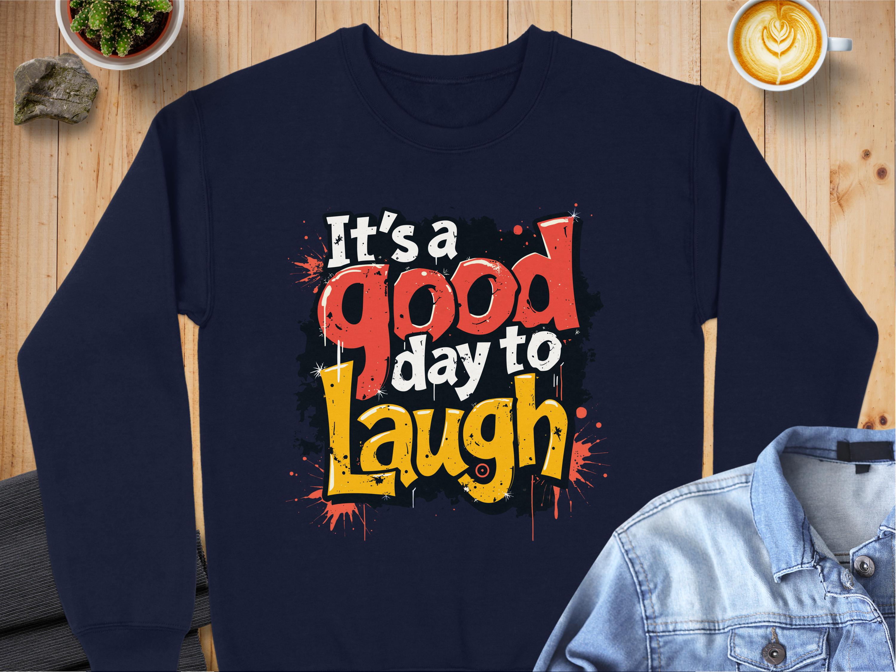 Garment Graphics offers a navy sweatshirt with Its a good day to Laugh in vibrant letters, featuring unisex sizing and medium-heavy fabric. Its styled with a denim jacket, a latte, and a small plant on wood.