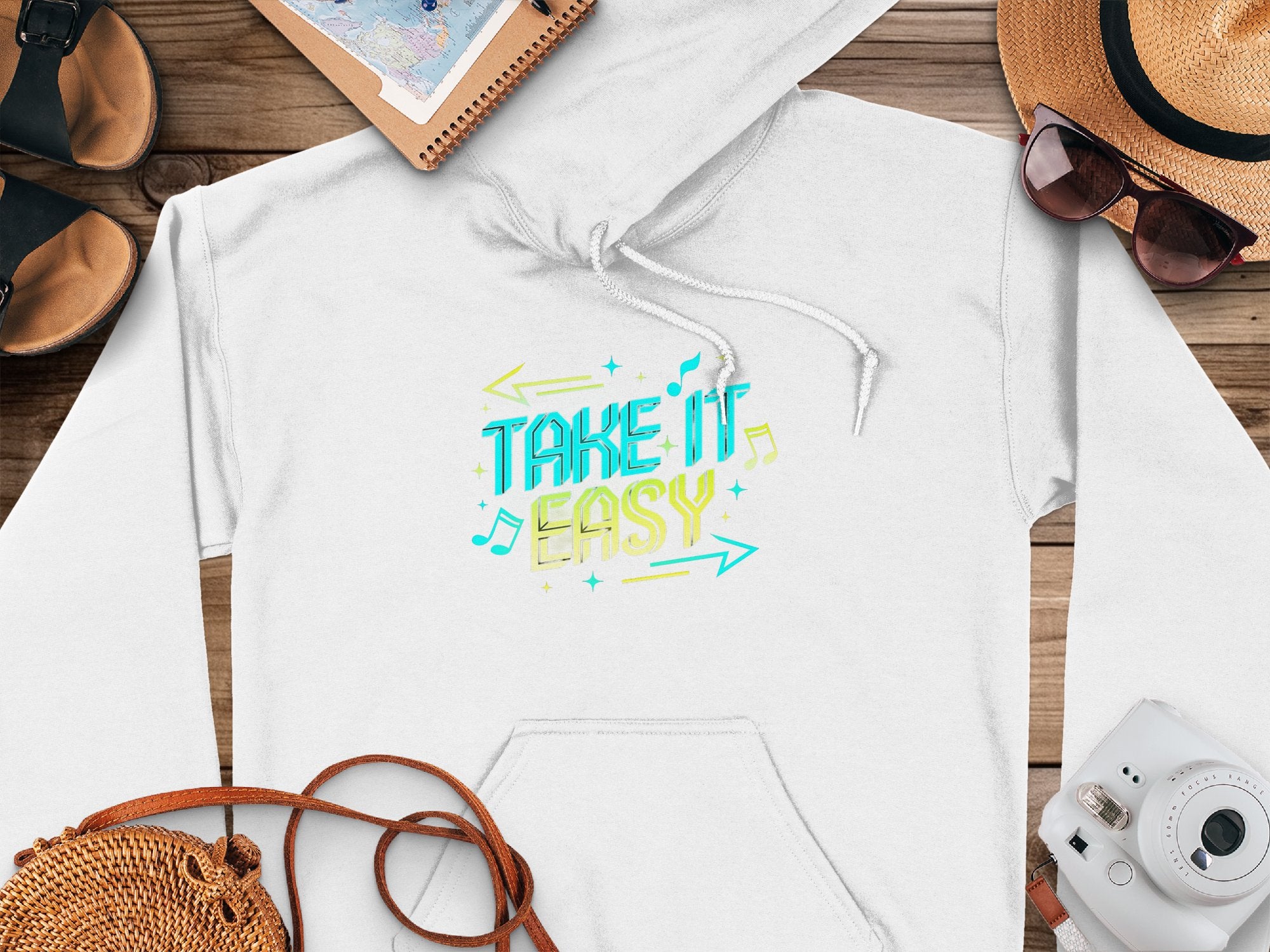 Garment Graphics presents a white hoodie with Take It Easy in vivid blue and yellow. Laid on wood, its accompanied by sunglasses, a sun hat, sandals, a notebook, pen, and camera—embodying relaxed fashion and casual style.