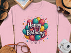 A pink Garment Graphics sweatshirt featuring vibrant balloons and Happy Birthday text is showcased on a wooden surface alongside a straw hat, sunglasses, a camera, sandals, and a notebook. This celebratory unisex sweatshirt is available for everyone to enjoy.