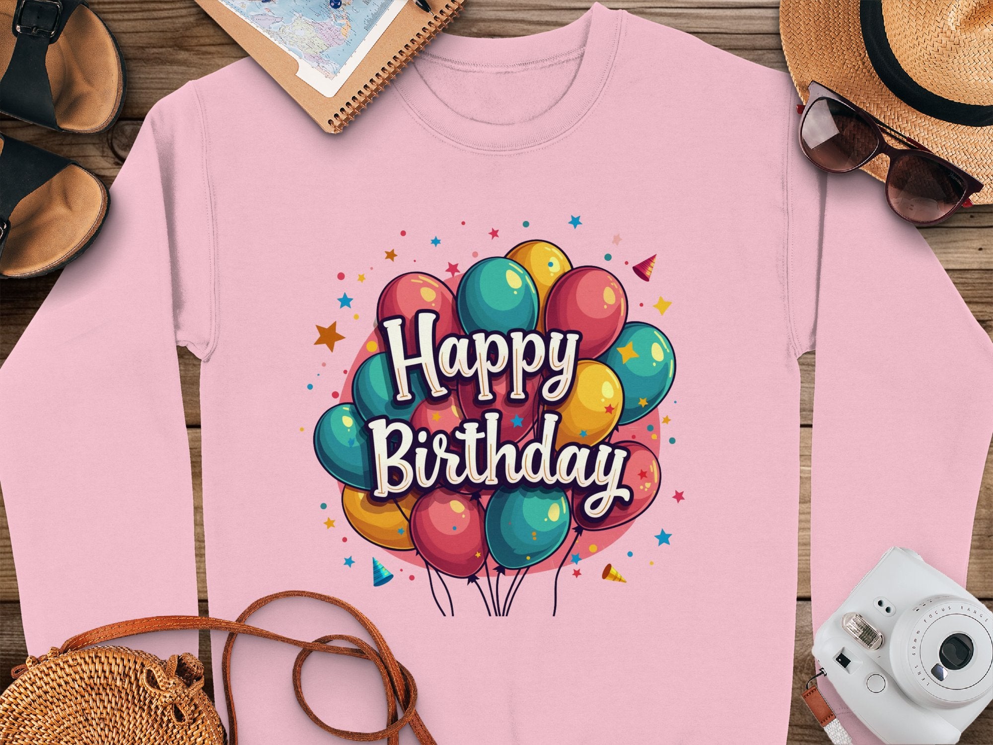 A pink Garment Graphics sweatshirt featuring vibrant balloons and Happy Birthday text is showcased on a wooden surface alongside a straw hat, sunglasses, a camera, sandals, and a notebook. This celebratory unisex sweatshirt is available for everyone to enjoy.
