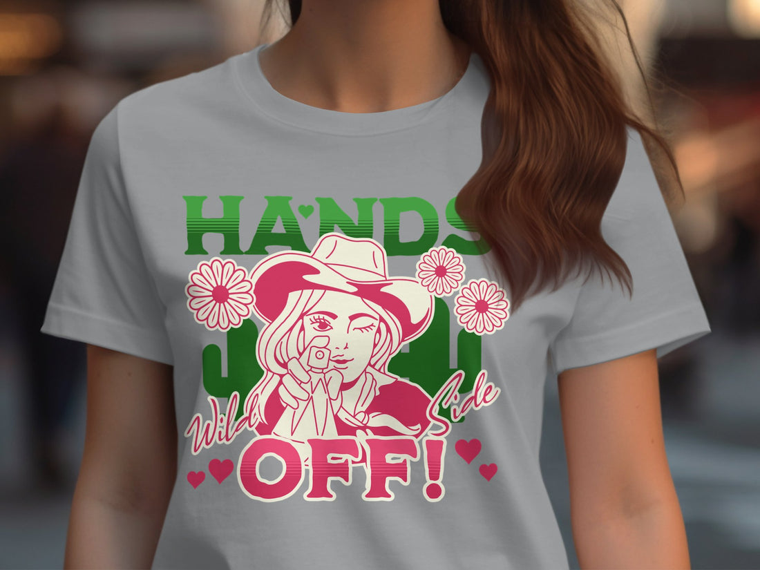 This bold Garment Graphics cowboy T-shirt showcases a person in a cowboy hat surrounded by daisies with Hands Off! in vibrant green and red. Set on wood, its framed by Christmas decorations, a cozy sweater, and wrapped gifts for a unique holiday style.