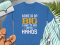 A vibrant blue sweatshirt by Garment Graphics boldly says, MINE IS SO BIG I HAVE TO USE BOTH HANDS. It sits amidst a striped shirt, brown sandals, a wristwatch, and a green bag on ferns against a gray backdrop.